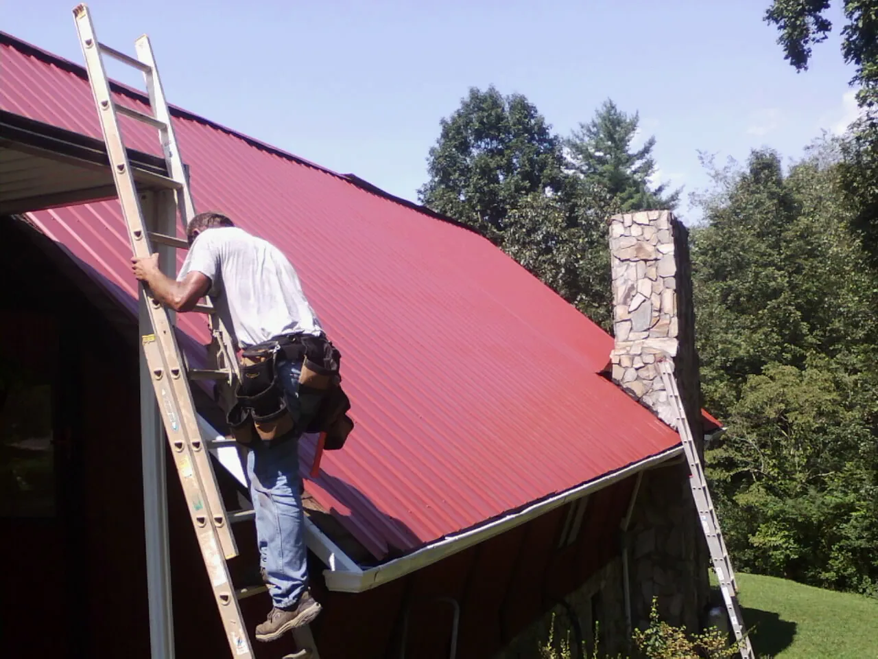 Roofer on a ladder beside a red metal roof and stone chimney — Soffit & Fascia Repair by Roofing Brothers in Bandon